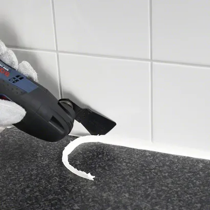 Gloved hand uses a multitool to remove silicone sealant from a tiled surface.