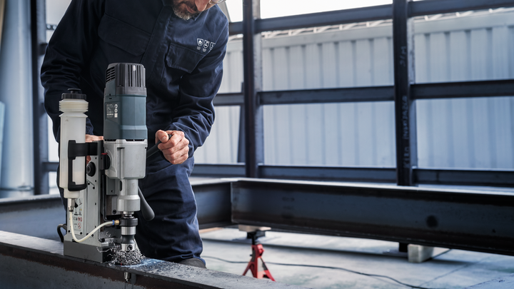 A person wearing safety equipment operates a magnetic drill on a steel beam.