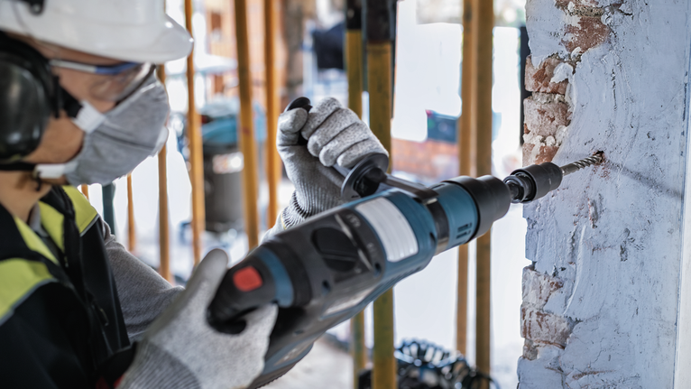 A person wearing safety equipment drills into a concrete wall.