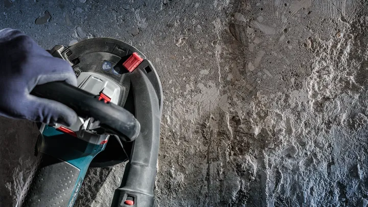 Person wearing safety equipment operates a power tool on a rough concrete wall.