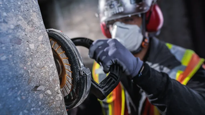 Person wearing safety equipment grinds concrete with a power tool.