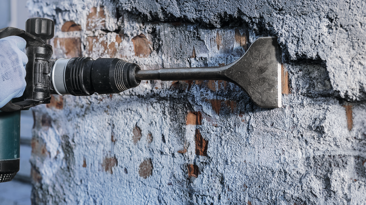 Person wearing safety equipment uses a chisel attachment to remove plaster from a wall.