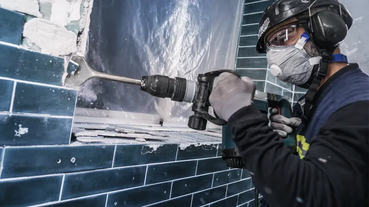 A person wearing safety equipment removes tiles with a demolition hammer.