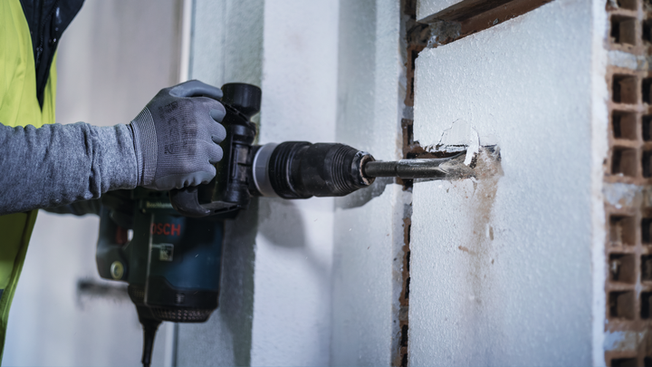 A person wearing safety equipment uses a power drill to bore a hole in a wall.