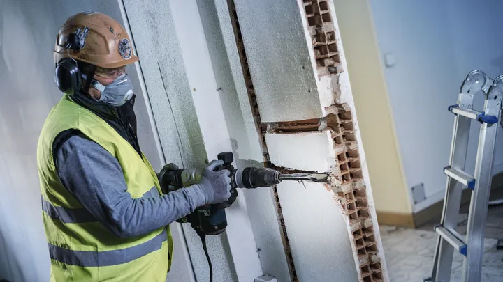 Person wearing safety equipment drills into a wall in a construction area.