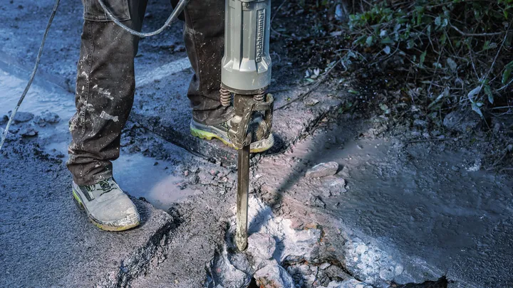 Person wearing safety equipment breaking pavement with a demolition tool.
