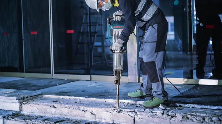 A person wearing safety equipment uses a demolition hammer to break concrete.