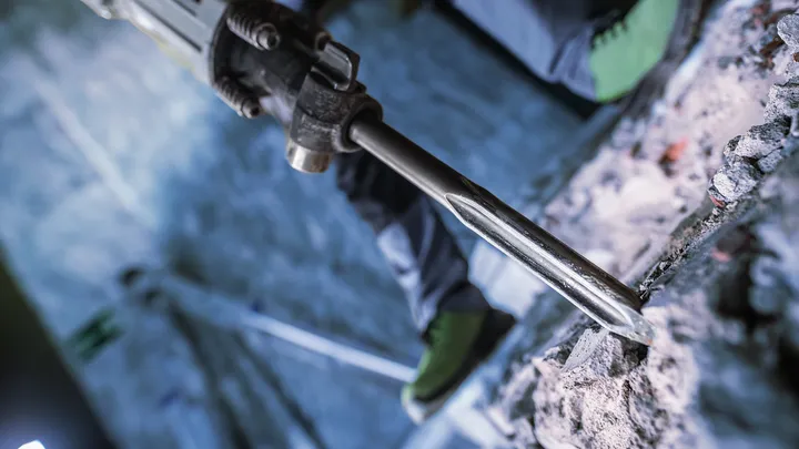 Person wearing safety equipment uses a demolition hammer to break concrete.
