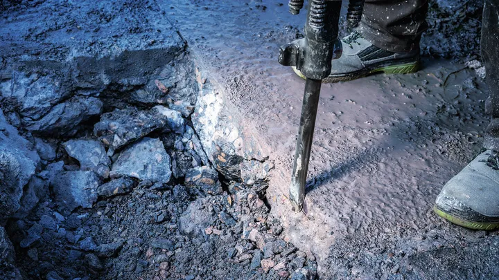 A person wearing safety equipment uses a demolition tool to break concrete.