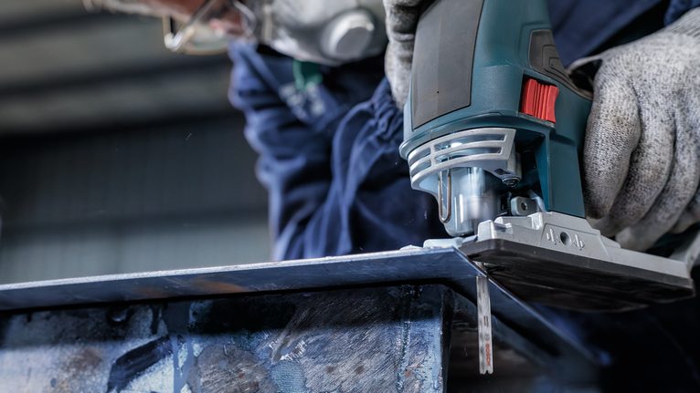 Person wearing safety equipment uses a jigsaw to cut through a metal sheet.