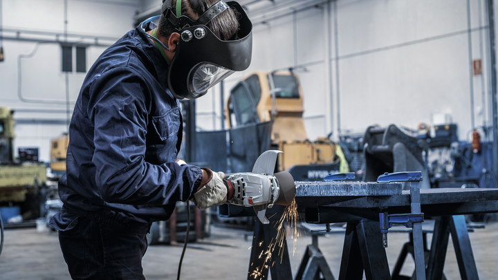 Person wearing safety equipment grinds metal, producing sparks in a workshop.