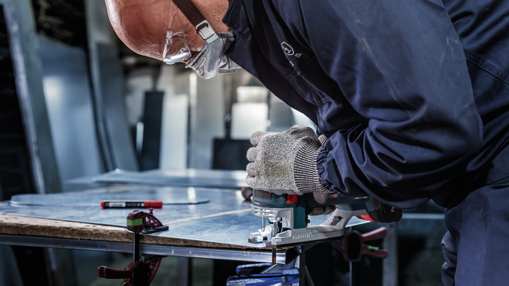 A person wearing safety equipment cuts sheet metal with a jigsaw.