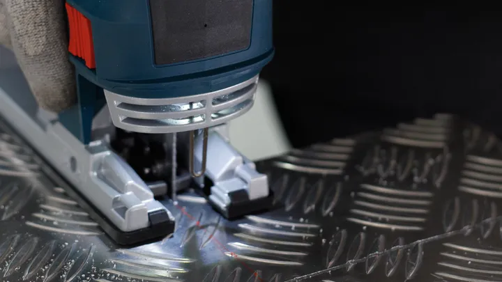Person wearing safety equipment operates a jigsaw to cut a patterned metal sheet.