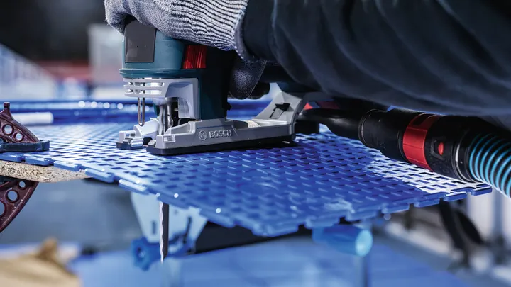 Person wearing safety equipment guides a jigsaw to cut a blue perforated sheet.
