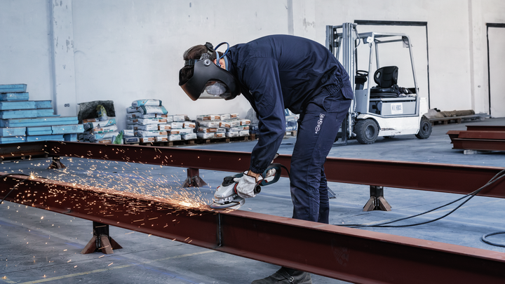 A person wearing safety equipment grinds a metal beam, producing sparks in a warehouse.
