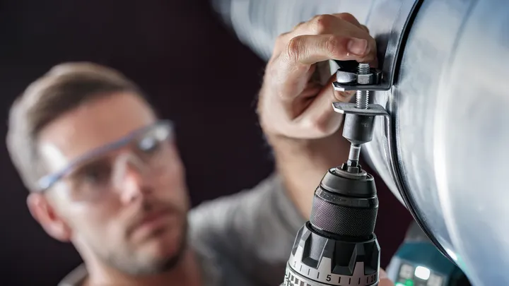 Person wearing safety equipment drills a hole into a large metal pipe.