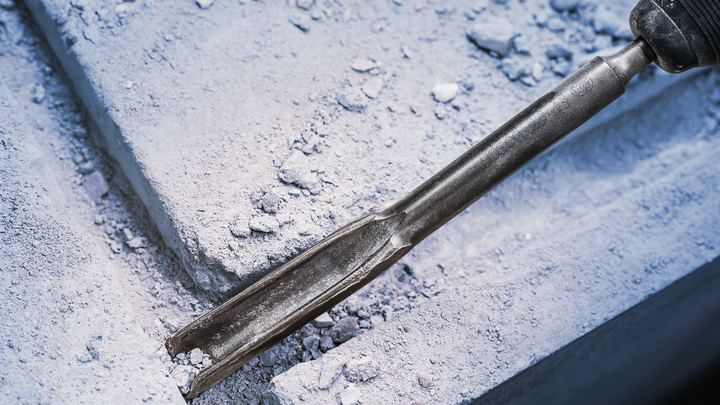 Chisel tool breaking concrete with dust and debris scattered around.