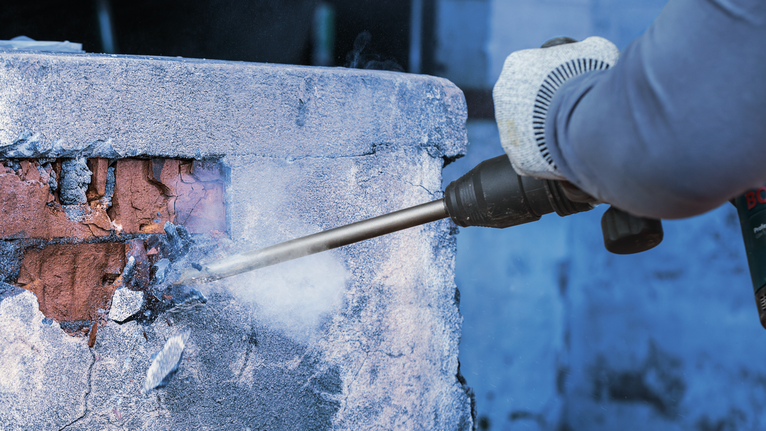 Person wearing safety equipment uses a power tool to break concrete on a wall.