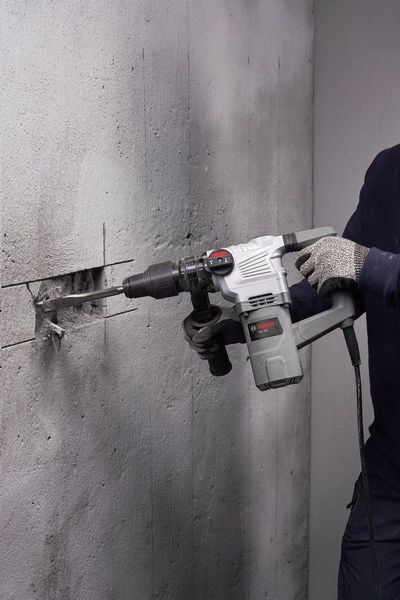 A person wearing safety equipment uses a rotary hammer to chisel a groove in a concrete wall.