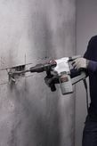 A person wearing safety equipment uses a rotary hammer to chisel a groove in a concrete wall.