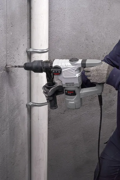 A person wearing safety equipment drills into a concrete wall with a rotary hammer.