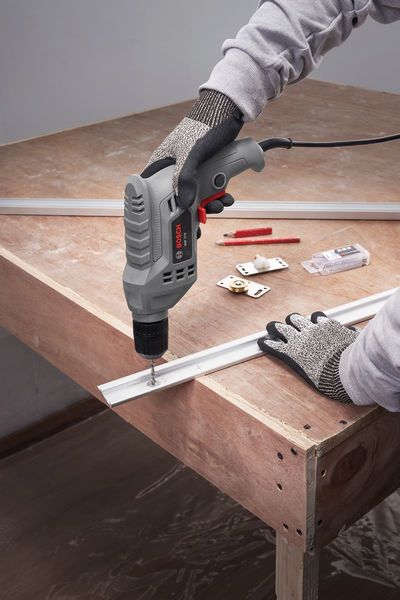 A person wearing safety equipment uses a drill to fasten a metal strip on a wooden table.