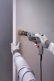 A person wearing safety equipment uses a drill to make a hole in a wall.