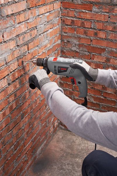 A person wearing safety equipment drills into a red brick wall with a power tool.