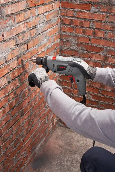 A person wearing safety equipment drills into a red brick wall with a power tool.