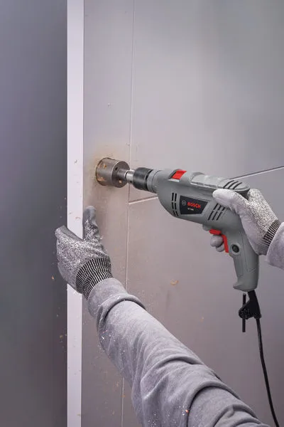 A person wearing safety equipment uses a power drill to bore a hole in a wall.