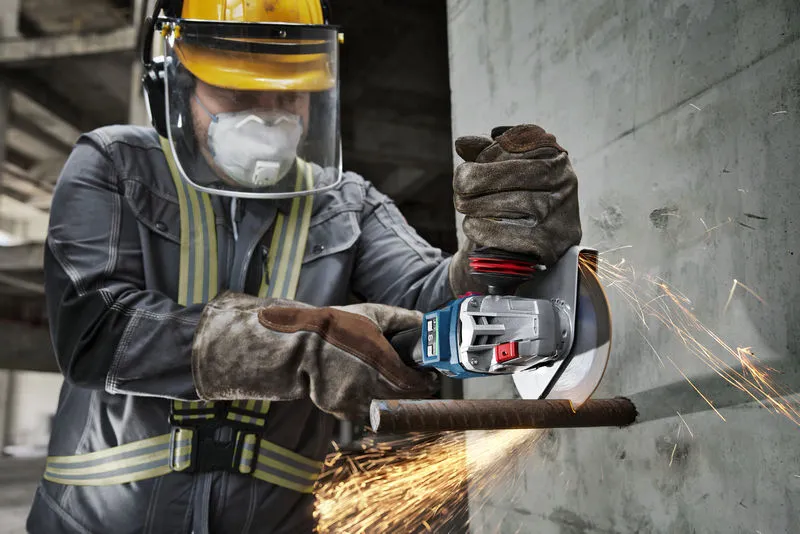 A person wearing safety equipment uses a cordless angle grinder to cut through rebar.