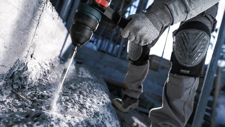 A person wearing safety equipment uses a rotary hammer to drill into concrete.