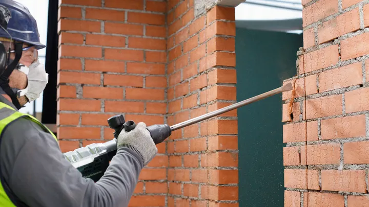 Person wearing safety equipment chisels a brick wall with a power tool.