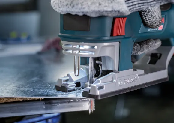 Person wearing safety equipment uses a jigsaw to cut a metal sheet.