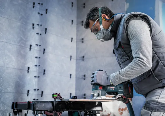 A person wearing safety equipment uses a jigsaw to cut wood on a workbench.