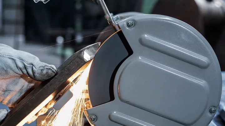 A person wearing safety equipment grinds a metal bar with a bench grinder.