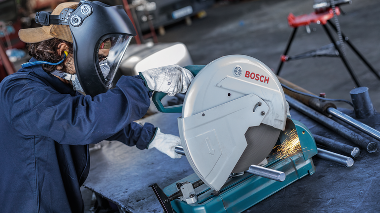 Person wearing safety equipment cuts metal pipes with a circular saw, sparks flying.