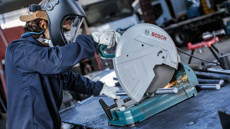 Person wearing safety equipment cuts metal pipes with a power saw, creating sparks.