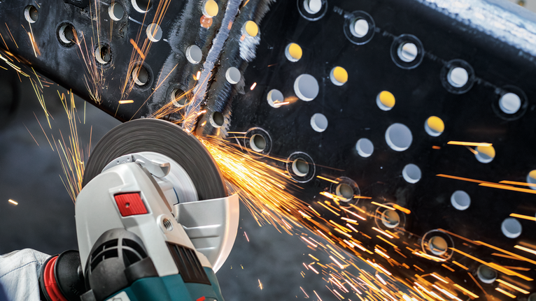 Person wearing safety equipment grinds a metal beam with a power tool, creating sparks.