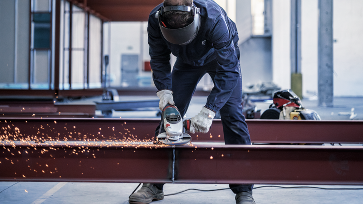 Person wearing safety equipment grinds a steel beam, producing bright sparks.