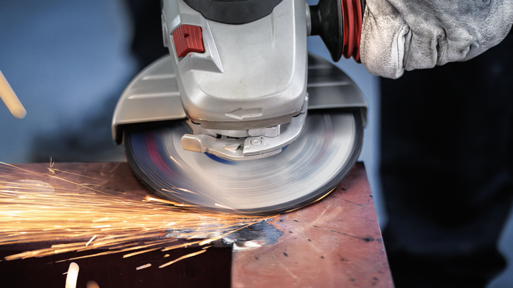 Person wearing safety equipment grinds metal using an angle grinder, creating sparks.