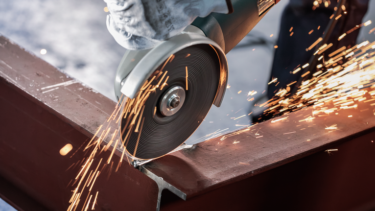 Person wearing safety equipment uses an angle grinder to cut a steel beam.