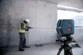 A person wearing safety equipment uses a drill while a laser leveling tool projects a red line.