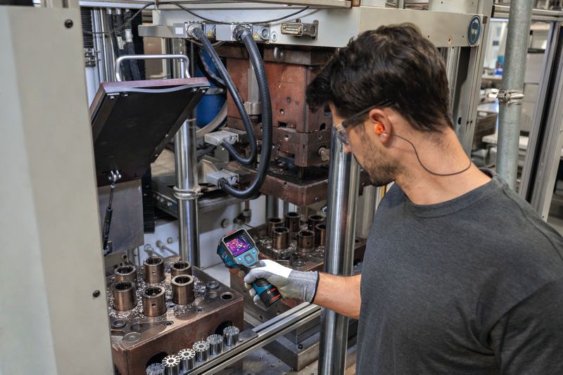 A person wearing safety equipment inspects industrial machinery with a thermal camera.