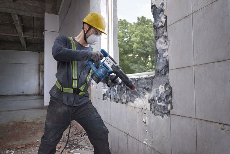 A worker wearing safety equipment uses a demolition hammer to break a tiled wall.