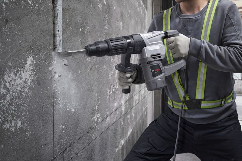 A person wearing safety equipment uses a demolition hammer to break concrete wall.