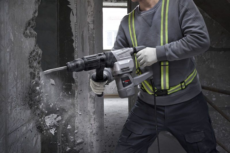 A person wearing safety equipment uses a demolition hammer to break a concrete wall.