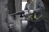 A person wearing safety equipment uses a demolition hammer to break a concrete wall.
