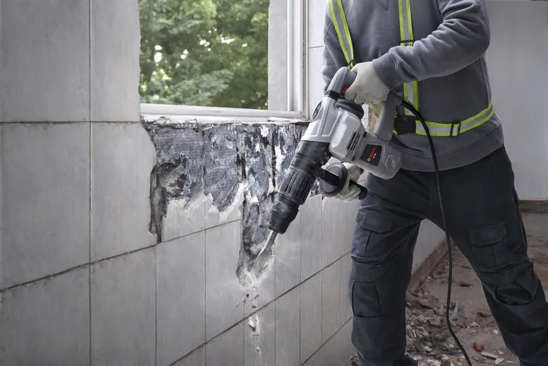 A person wearing safety equipment uses a demolition breaker to remove wall tiles.