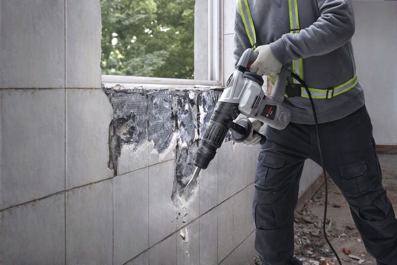 A person wearing safety equipment uses a demolition breaker to remove wall tiles.